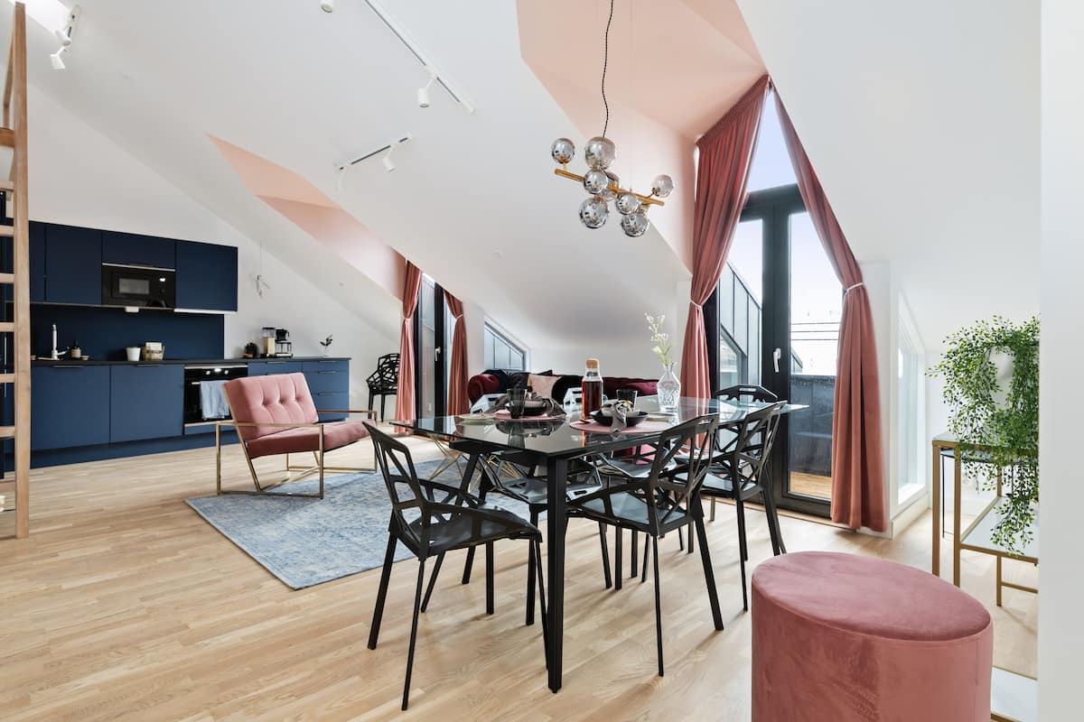 Una cocina moderna con armarios oscuros y un microondas, un comedor con una mesa de vidrio y sillas negras, y un banco con cojín rojo.