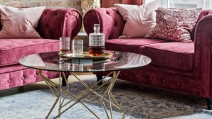 A room with a brick wall, a pink sofa, a round glass coffee table with metal legs, and a window with red curtains.