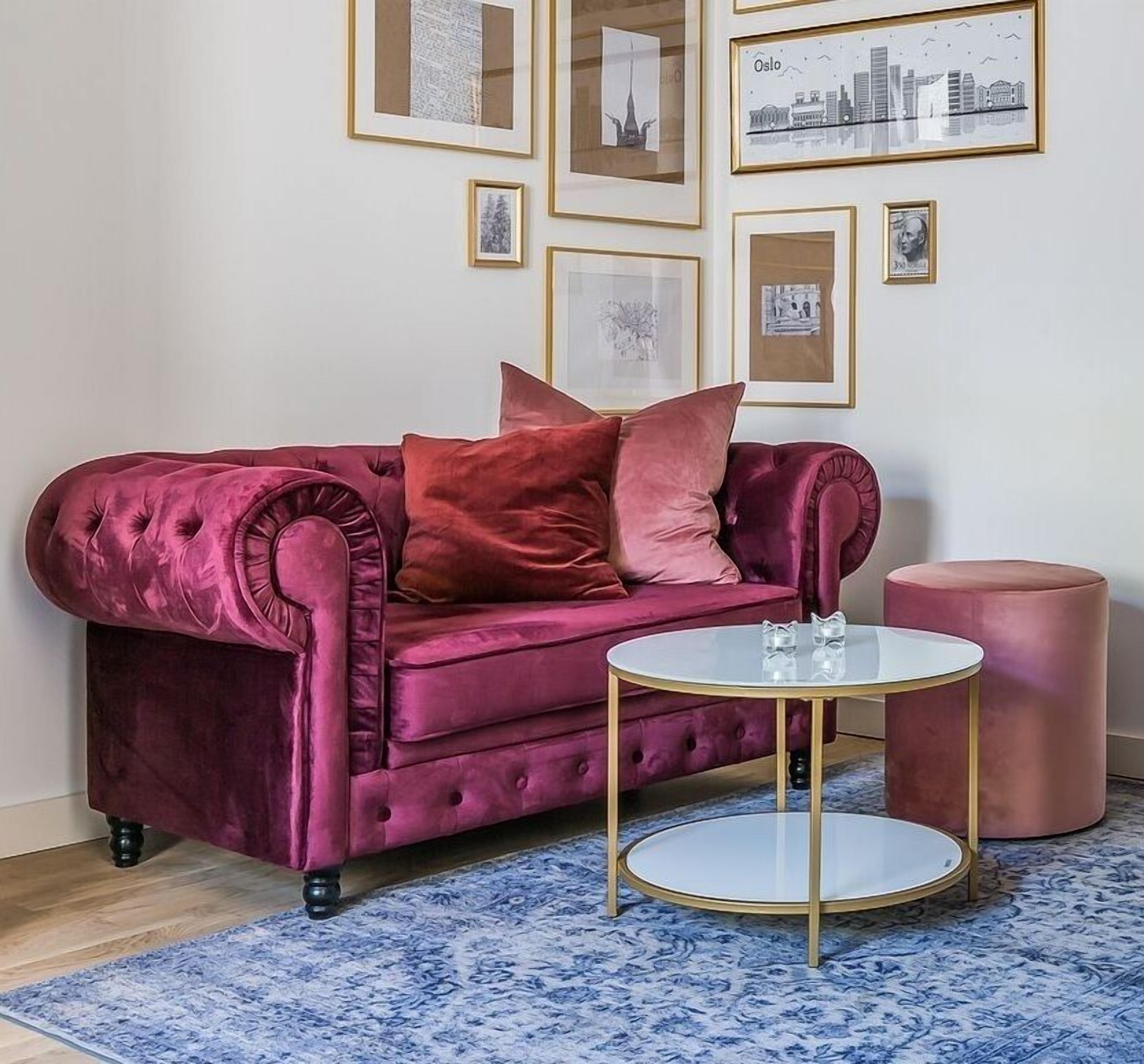 Un canapé moelleux en velours rouge, doté d’un dossier et de bras capitonnés, une table basse ronde en verre avec des pieds dorés, et un pouf assorti. La pièce est agrémentée d’un tapis bleu à motifs et d’œuvres d’art encadrées accrochées au mur.
