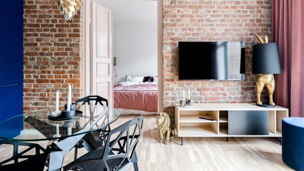 A modern living room with a brick wall, a TV mounted on the wall, a glass dining table, and a sofa.