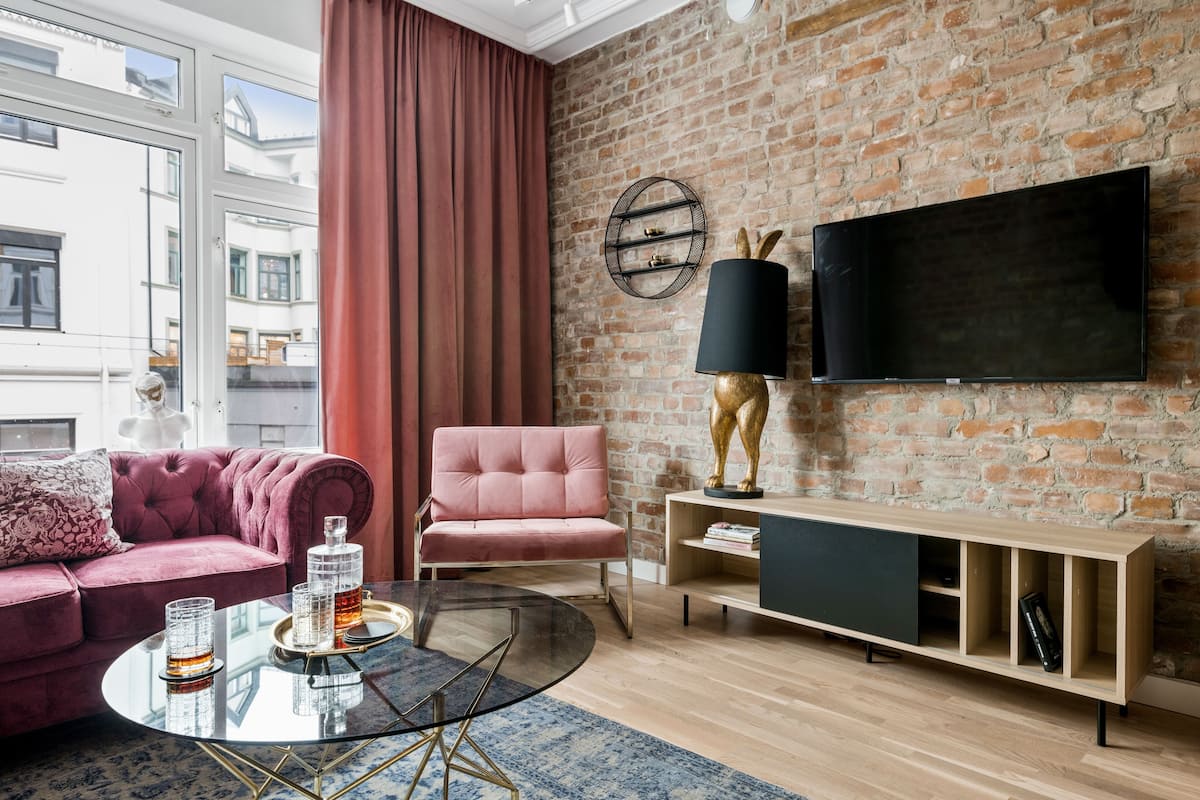 Una sala de estar moderna con una pared de ladrillo, un sofá rosa, una mesa de centro redonda de vidrio y un televisor montado.