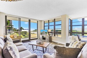 A spacious living room with a large sofa set, a coffee table, and a TV on a stand. The room has large windows offering a view of the outdoors.