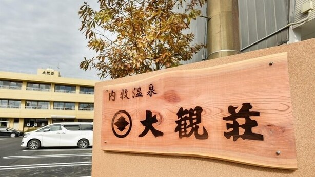 A wooden sign with Japanese characters, a parking lot, and a building in the background. - Aso Uchinomaki Onsen Daikanso (Aso)