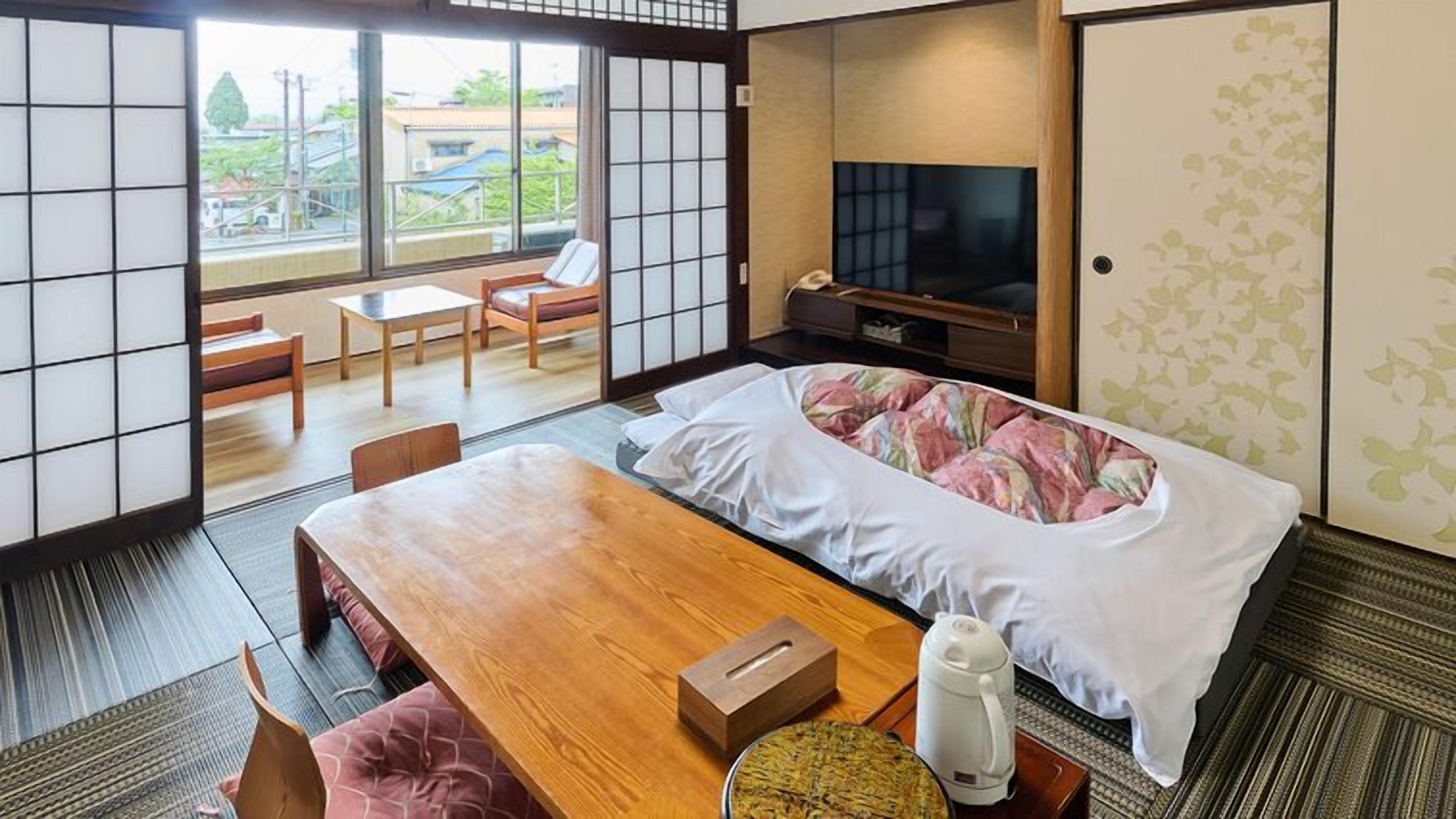 A traditional Japanese-style room with tatami flooring, a low wooden table, a bed with a floral bedspread, a television, and a sliding door with a view of the outside.