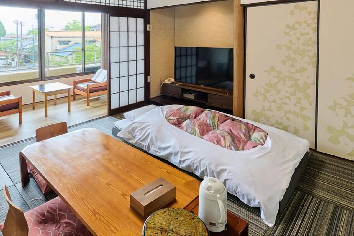 Una habitación tradicional japonesa con suelo de tatami, una mesa de madera baja, una cama con colcha estampada, un televisor y una puerta corredera con vistas al exterior.