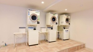 A laundry facility with four washing machines, each with a control panel and a basket on a small table. - Aso Uchinomaki Onsen Daikanso (Aso)
