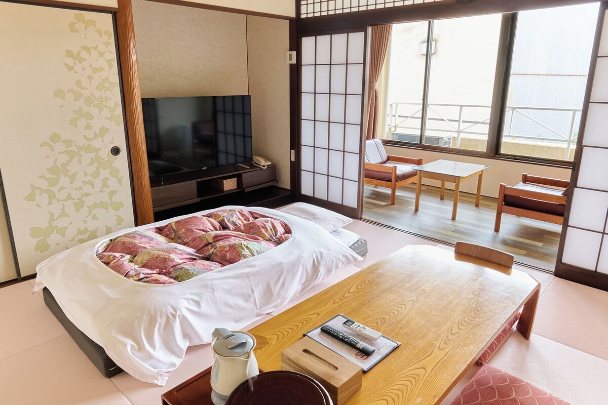 Une pièce de style japonais traditionnel, avec un sol recouvert de tatamis, un futon servant de lit, une télévision et une porte coulissante donnant sur un balcon.