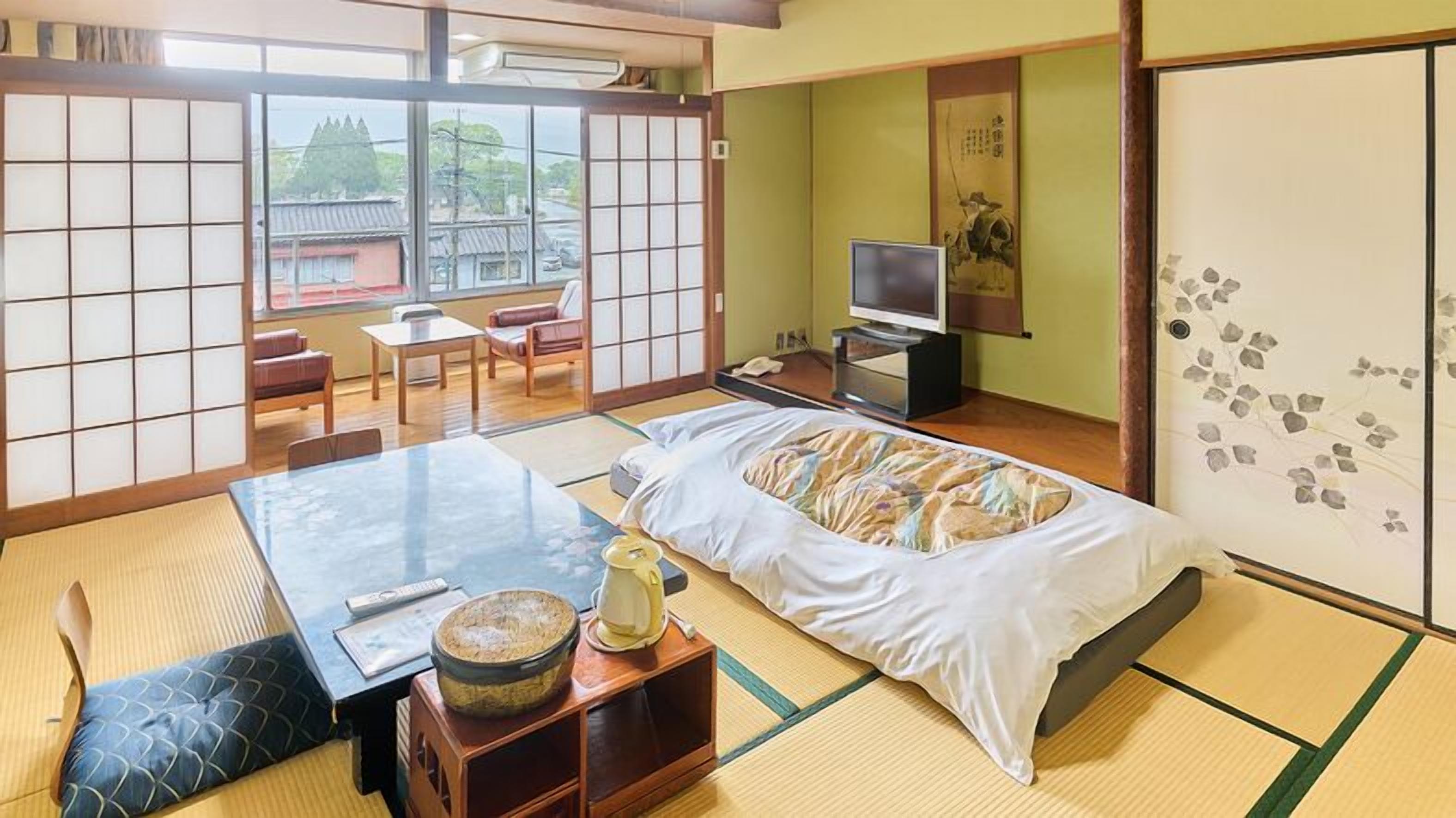A traditional Japanese room with tatami flooring, a bed with a colorful blanket, a television, and a sliding door with a decorative pattern.