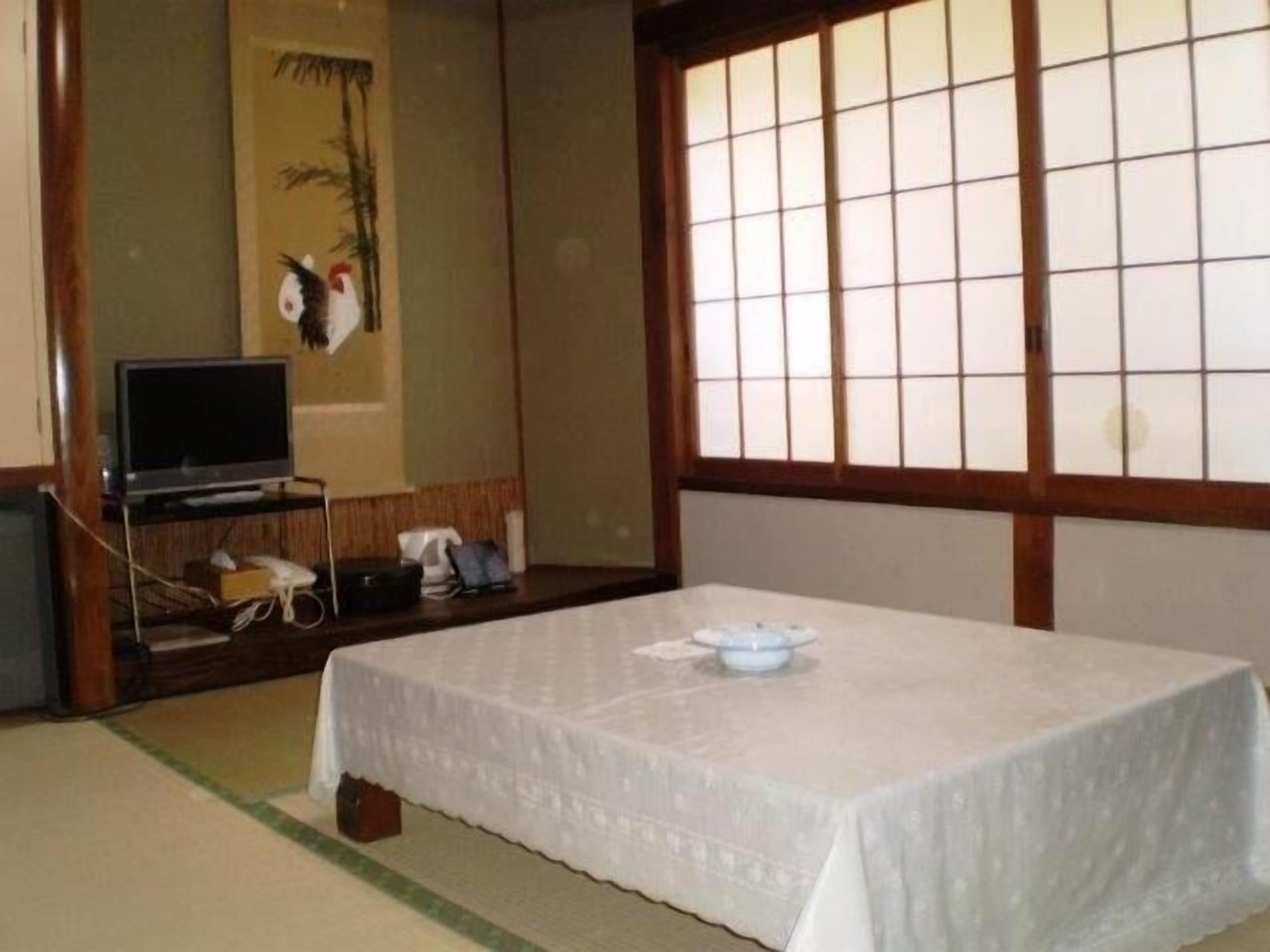 Una habitación japonesa tradicional con suelo de tatami, una mesa baja de tatami con mantel blanco, un televisor sobre un soporte de madera y una ventana con celosías shoji.