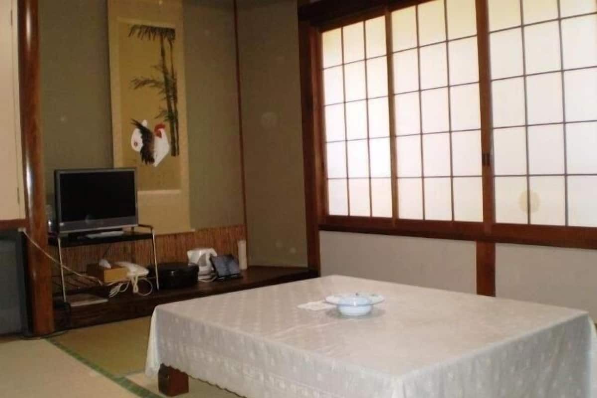 Une pièce de style japonais traditionnel, avec un sol recouvert de tatamis, une table basse en tatami recouverte d’une nappe blanche, un téléviseur posé sur un meuble en bois et une fenêtre à panneaux coulissants de type shoji.