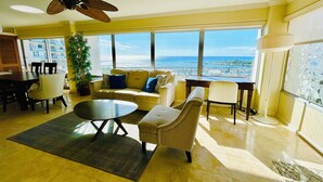 Un amplio salón con un gran ventanal con vistas a la playa, un ventilador de techo y un comedor con mesa y sillas.