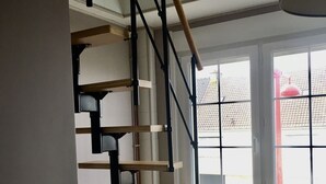 Una escalera de caracol con escalones de madera y barandillas de metal negro en una habitación con un radiador, una ventana que ofrece vistas a un edificio rojo y un techo blanco.