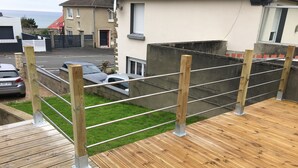 Una terraza de madera con barandas metálicas, un área con césped y vista a una calle residencial con casas y automóviles estacionados.