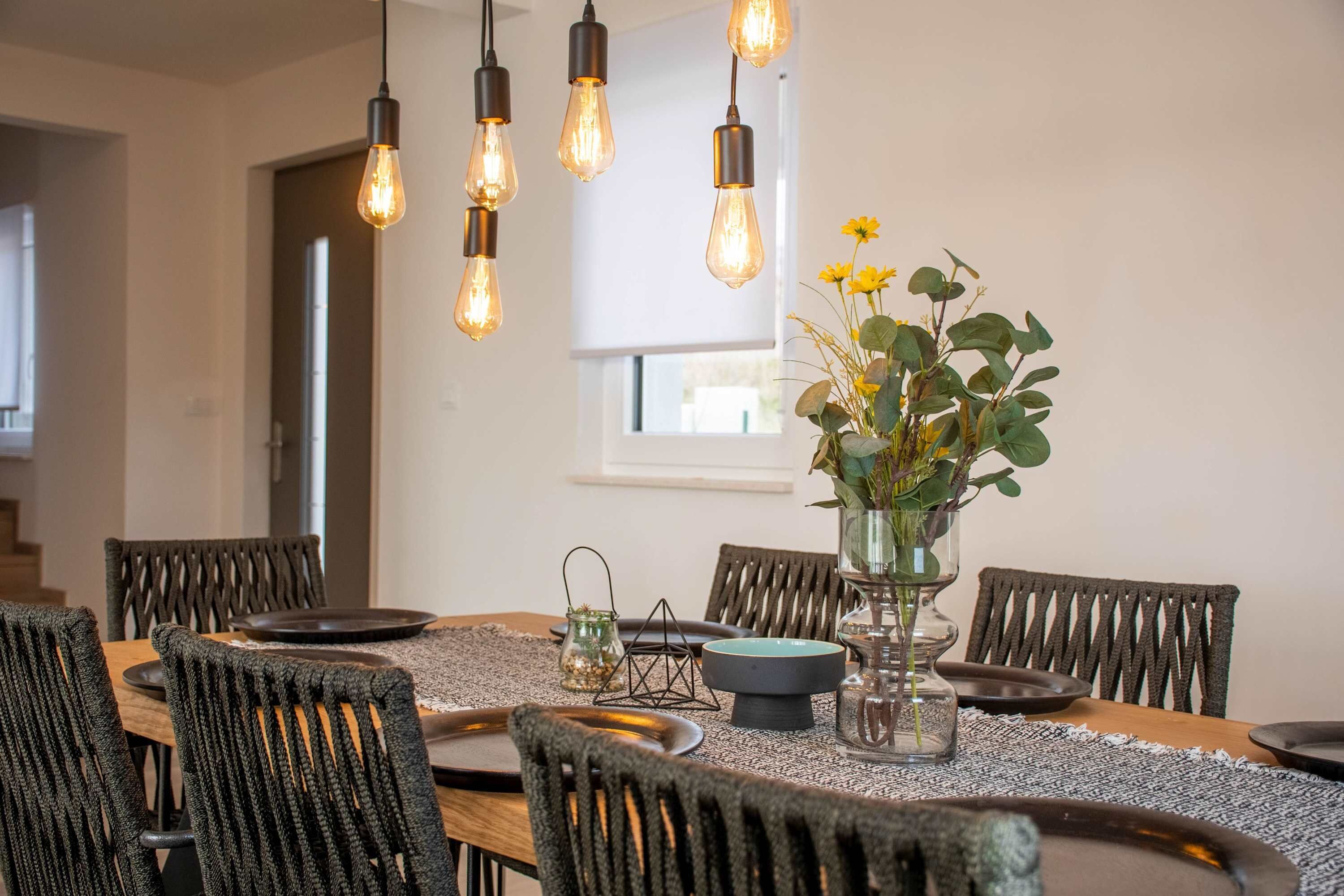 A dining area with a wooden table, woven chairs, hanging light bulbs, and a vase with flowers.