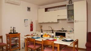 A compact kitchen with white cabinets, a gas stove, and a sink. There are chairs around a table with a bowl of fruit and bottles. - Tommy House - Convenient, functional and comfortable home (Orosei)