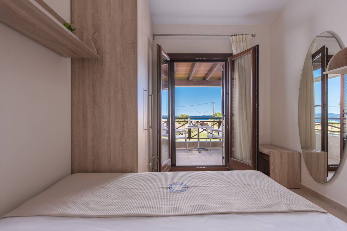 Ein Schlafzimmer mit einem Bett, einem Holzschrank, einem Spiegel und Blick auf eine Terrasse mit Tisch und Stühlen.