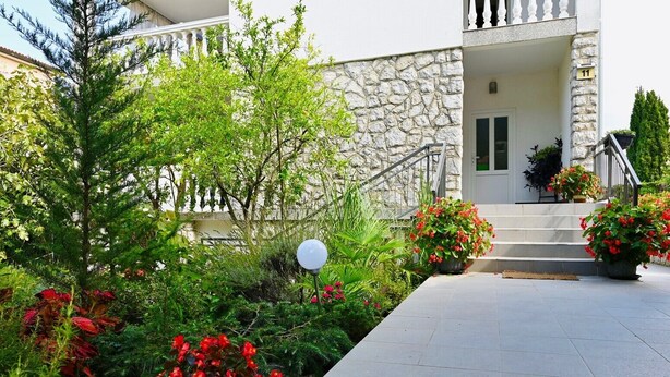 Un edificio de piedra con fachada blanca, una escalera que conduce a la entrada y un jardín bien cuidado con flores rojas y arbustos verdes.