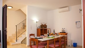A dining area with a table set for six, surrounded by wooden chairs with cushioned seats. A staircase with a metal railing is visible to the left, and an air conditioning unit is mounted on the wall to the right. - Casa Cristina - Convenient, functional and comfortable house (Orosei)