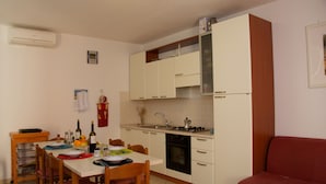 A compact kitchen with white cabinets, a stove, and a sink. There is a dining table with chairs and a red sofa in the living area. - Casa Cristina - Convenient, functional and comfortable house (Orosei)