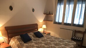 A bedroom with a wooden bed, two pillows, a nightstand with a lamp, a chair, a window with curtains, and a radiator. - Small villa in a great location (Viareggio)