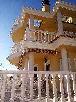 Un immeuble à plusieurs étages, doté d’une façade jaune, de balcons blancs et d’une terrasse carrelée avec des chaises blanches.