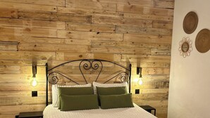 A bedroom with a wooden wall panel, a bed with white bedding and green pillows, two bedside tables, and a wall-mounted light fixture.