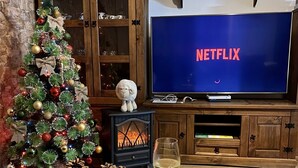 A cozy living room with a Christmas tree, a fireplace, a television displaying the Netflix logo, and a plate of crackers.