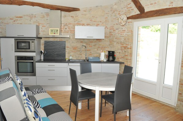Een keuken met een eettafel, stoelen, een magnetron, een spoelbak en een klok aan de muur.