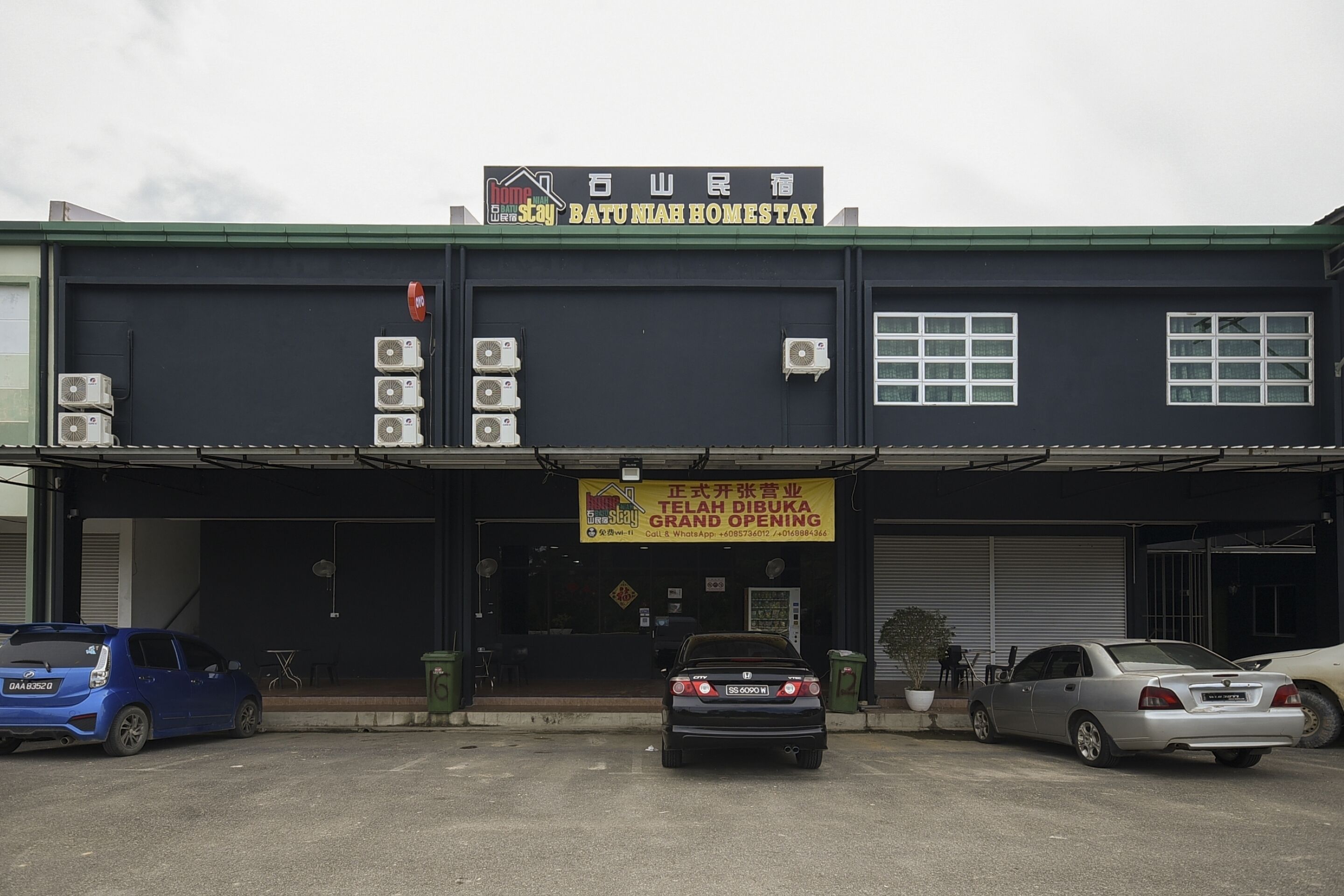 A building with a sign reading "Batu Mihah Homestay" and another sign indicating a grand opening. There are three cars parked in front.