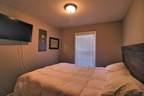 Une chambre à coucher comprenant un lit, un téléviseur fixé au mur, une fenêtre avec des stores et un luminaire au plafond.