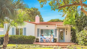 A two-story house with a red-tiled roof, a front porch with a bench, and a small garden with palm trees and shrubs.