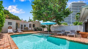 A swimming pool with a tiled deck, lounge chairs, and a large umbrella.