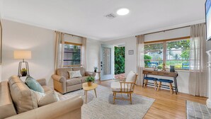 A living room with a sofa, armchair, and a small table with a lamp. There is a door leading to a balcony with a view of greenery.