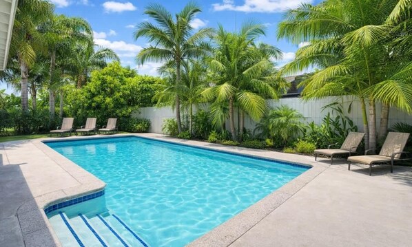 A rectangular swimming pool with clear blue water, surrounded by lounge chairs and palm trees.