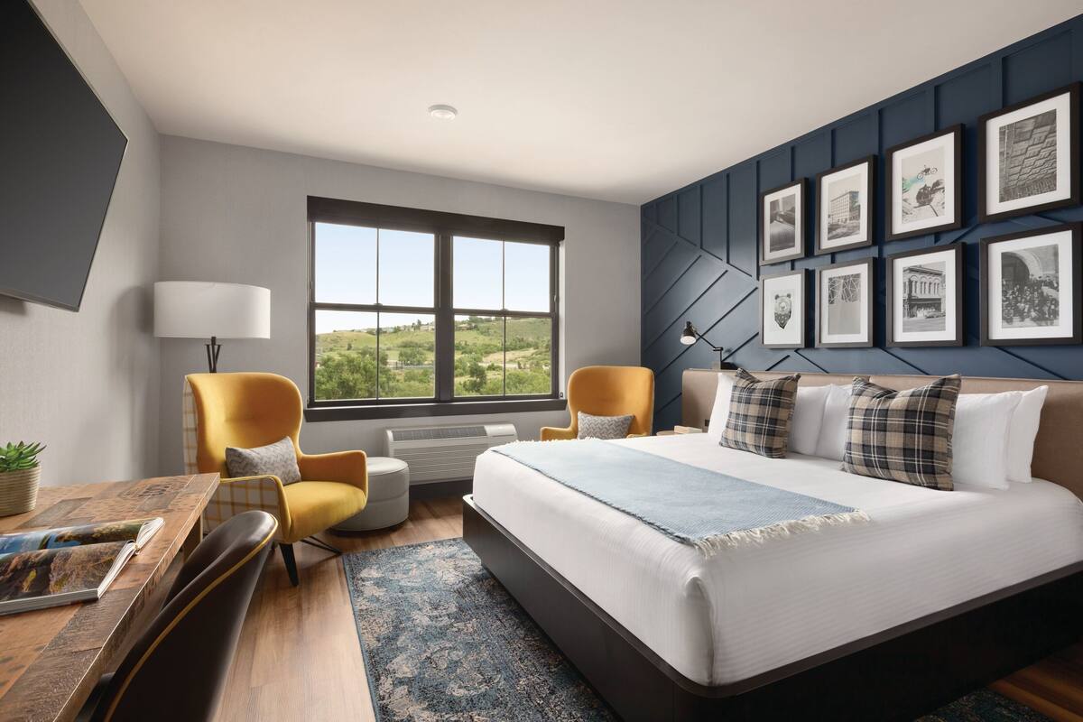 A modern hotel room with a large bed, two armchairs, a desk, and a large window offering a view of greenery.