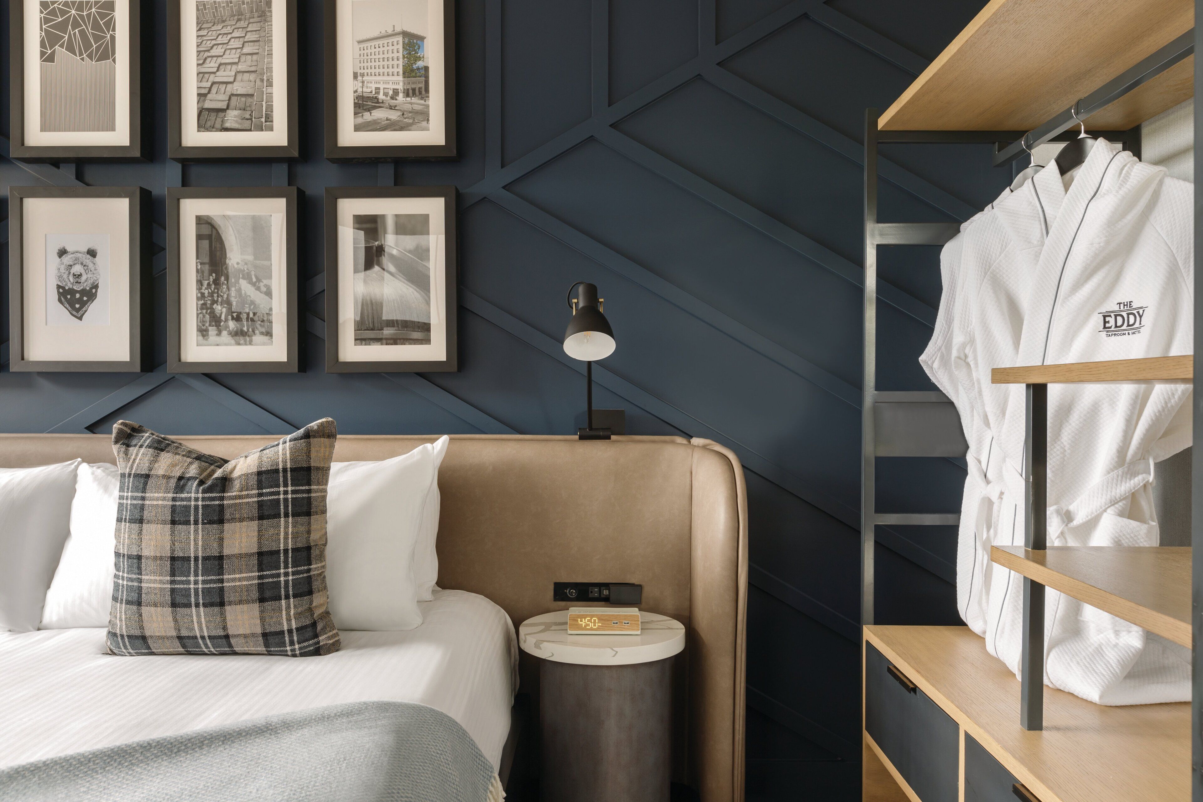 Een netjes ingerichte hotelkamer met een bed, een nachtkastje met een lamp, een klok en een plank met een badjas.