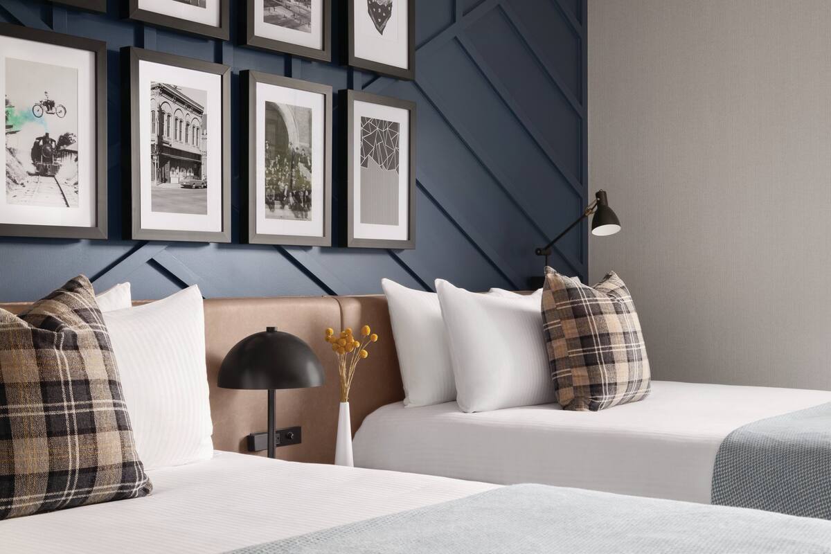A hotel room with two beds, a headboard, a bedside table with a lamp, and framed pictures on the wall.