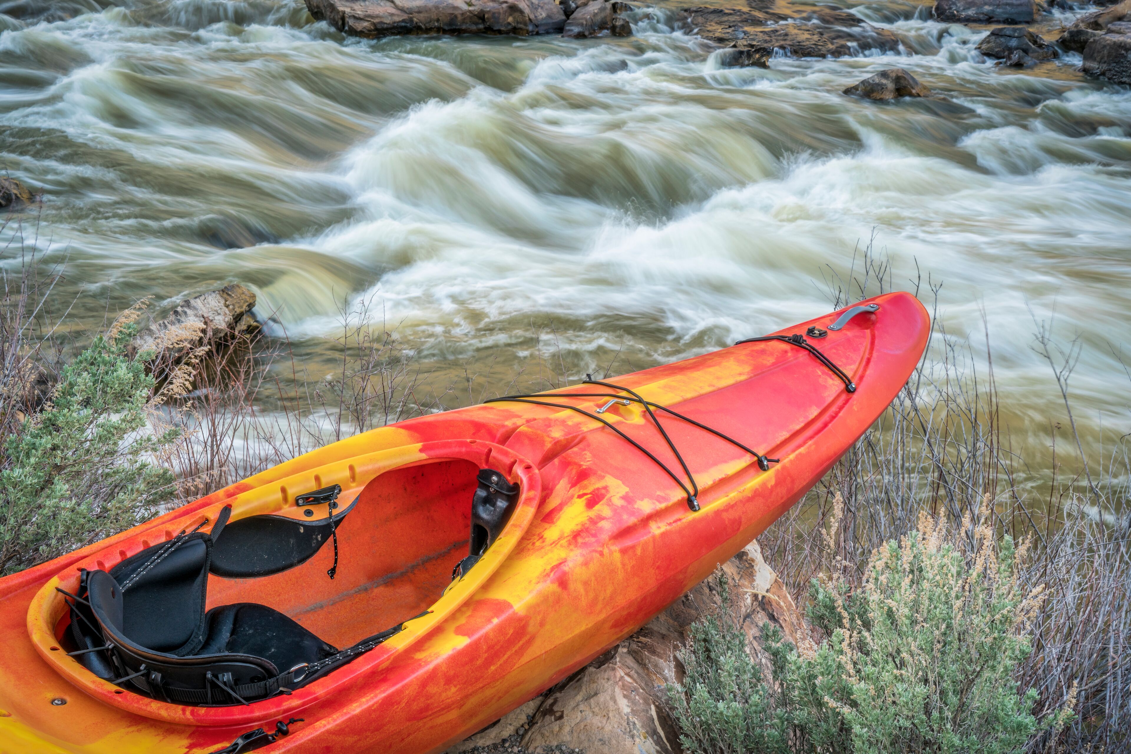 A red kayak with a black seat and black straps, resting on a rock by a flowing river.