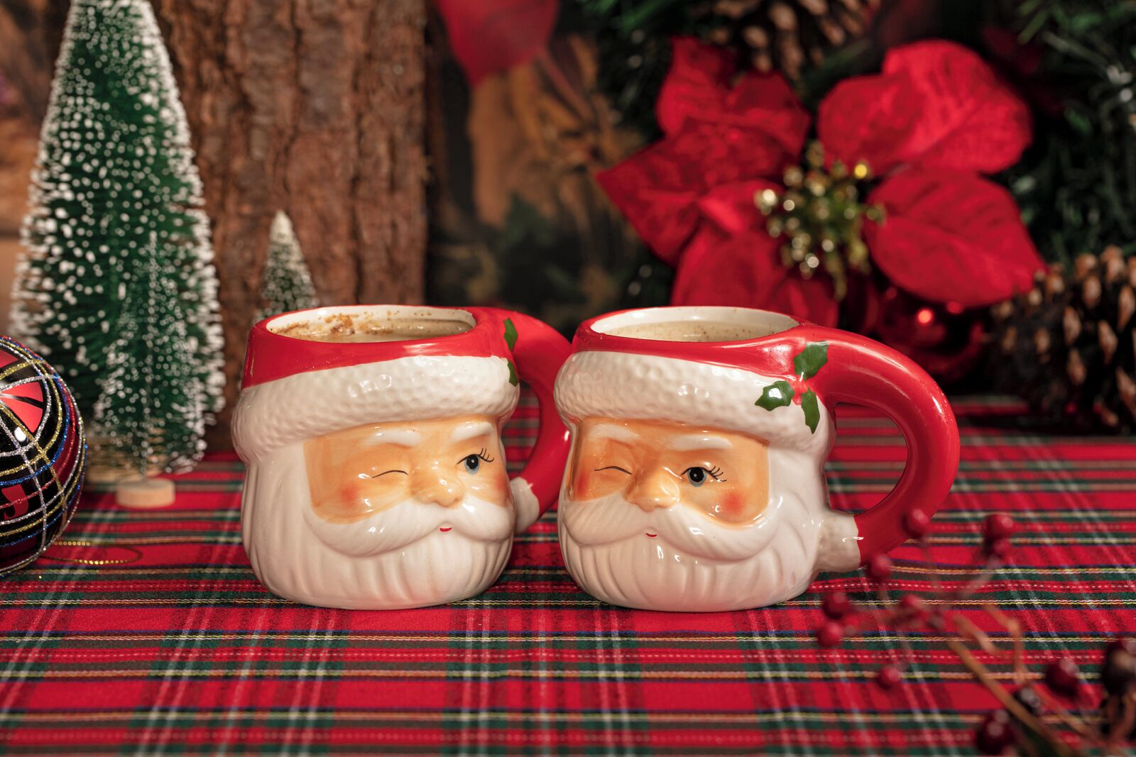 Twee keramikbekers met het figuur van de kerstman op een geruit tafelkleed, met een kerstboom en kerststerren in de achtergrond.