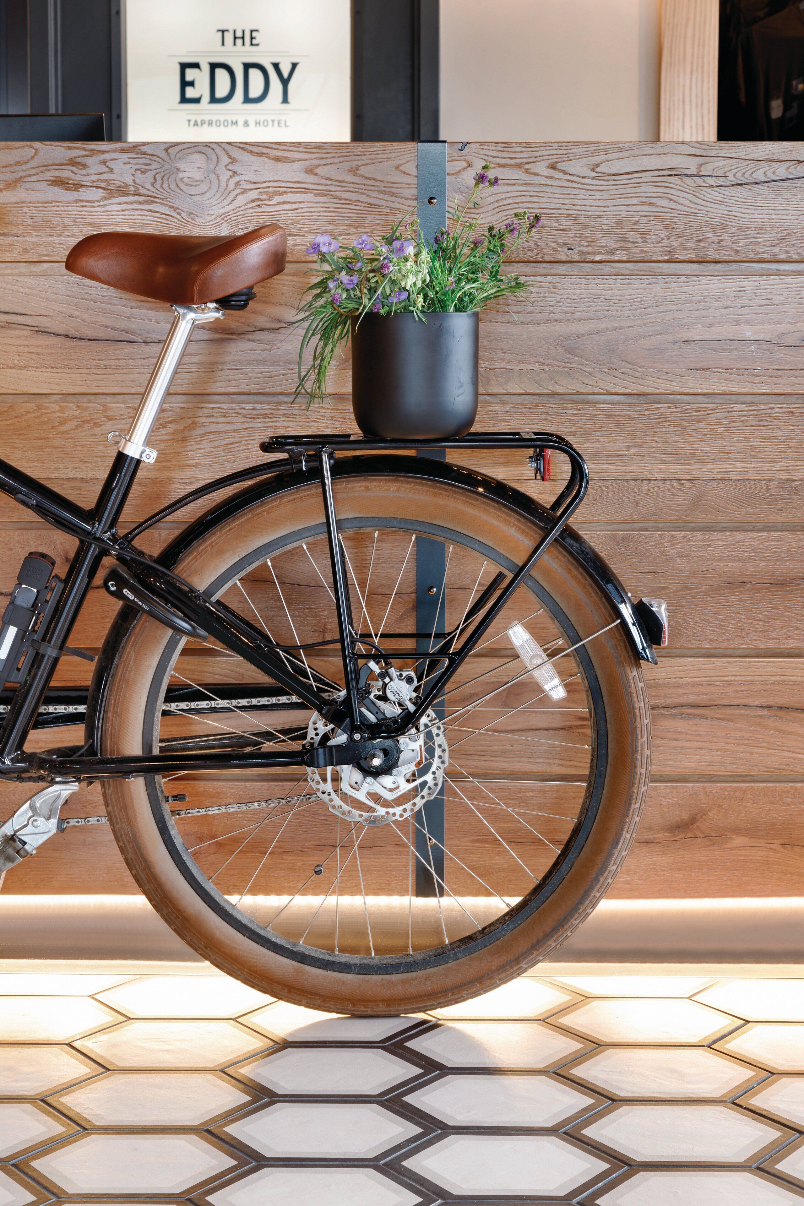 Una bicicleta con asiento de cuero y una planta en maceta sobre la rejilla trasera, estacionada frente a un muro de madera con un letrero que dice "THE EDDY TAPROOM & HOTEL".