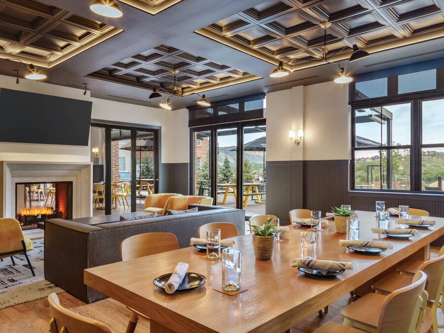 A dining area with a long wooden table set for a meal, a fireplace, and large windows offering a view of the outdoors.