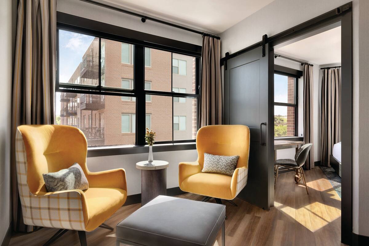 A modern hotel room with two yellow armchairs, a small round table, and a large window offering a view of the city.