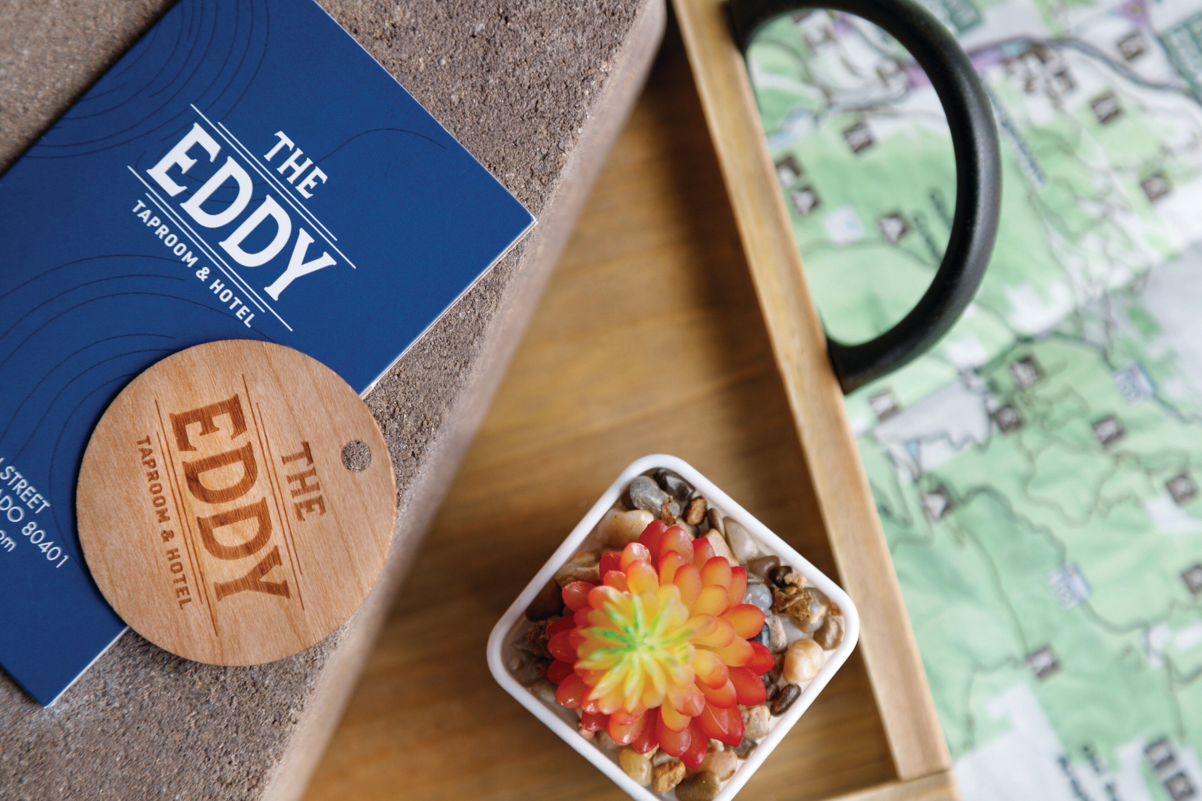 Una bandeja de madera con un mapa, un libro titulado "The EDDY TAPROOM & HOTEL" y un plato pequeño con un arreglo floral.