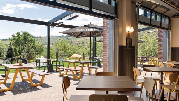 An outdoor patio with wooden tables and chairs, a large umbrella, and a view of the surrounding landscape. - The Eddy Taproom & Hotel (Golden)