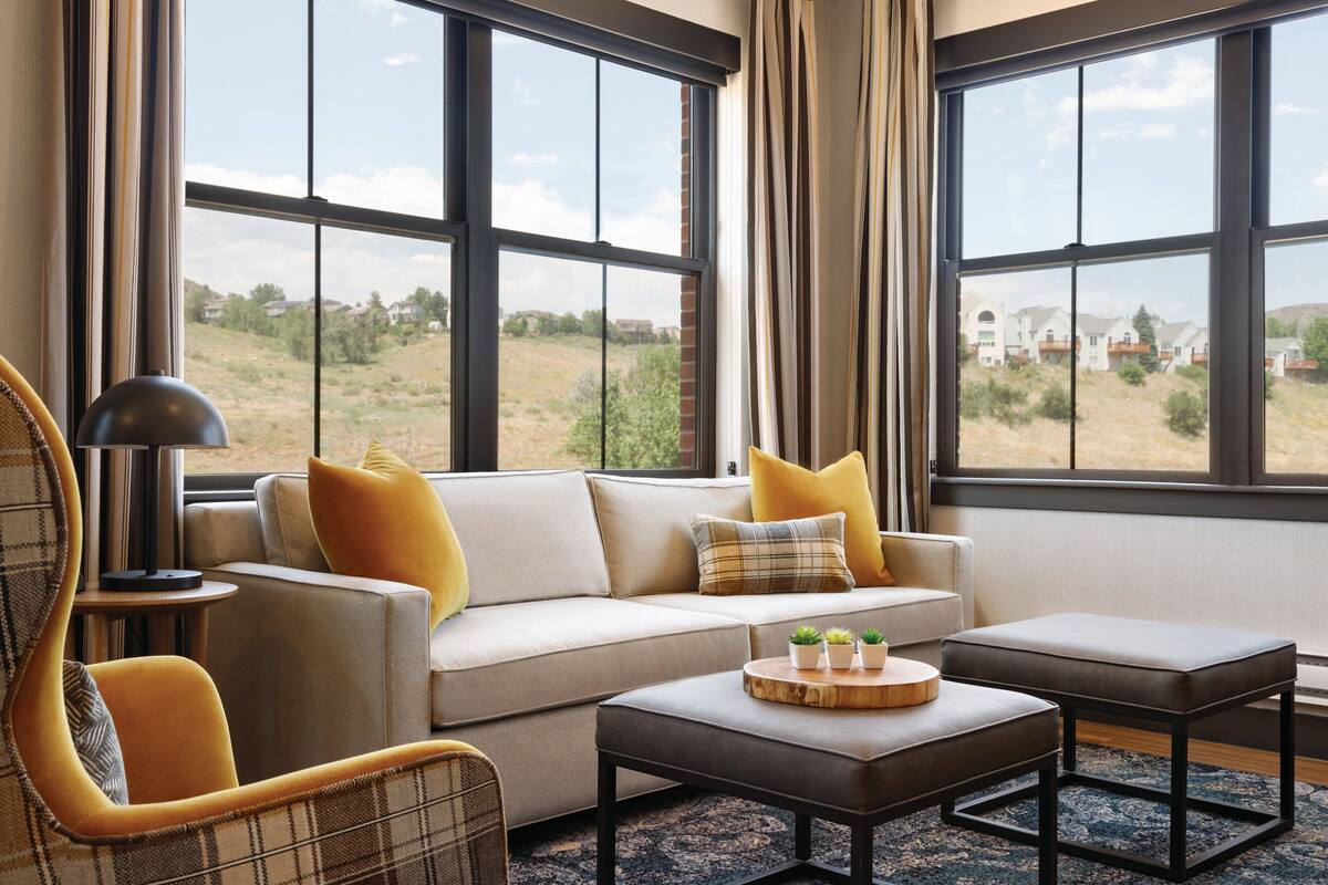 A modern living room with a sofa, armchair, ottoman, and a coffee table with plants. Large windows offer a view of the outdoors.