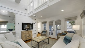 A spacious living room with a white sofa, a wooden coffee table, and a television. There is a balcony with wicker furniture and a view of the outside. - Renovated in Seaside, Sleeps 12! 3 Minutes to Beach! + 2 Adult Bikes! (Santa Rosa Beach)