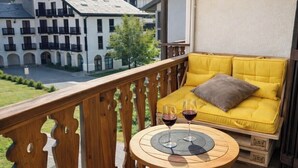 Un balcón con un sofá amarillo, dos copas de vino y vistas a un edificio con balcones.