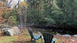 Zwei Adirondack-Stühle am Flussufer in einem bewaldeten Gebiet mit Steinen und herabgefallenen Blättern.