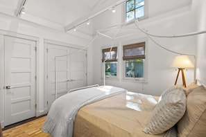 A bedroom with a bed, white walls, a window with blinds, a lamp on a tripod, and a ceiling light fixture. - Carmel's Wy Knot: Beautiful remodel in Carmel-by-the-Sea! (Carmel)