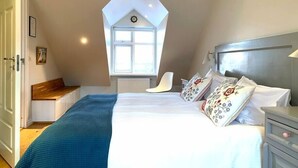Un dormitorio con techo inclinado, una cama grande con almohadas estampadas, una silla blanca y una ventana con vistas.